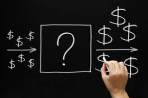 A hand is holding white chalk and drawing dollar signs on a blackboard to portray the idea of an investment.